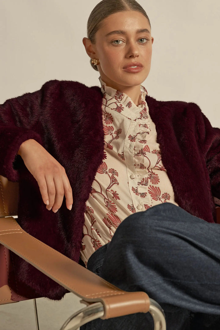 Woman wearing a floral blouse and dark coat sitting on a chair.
