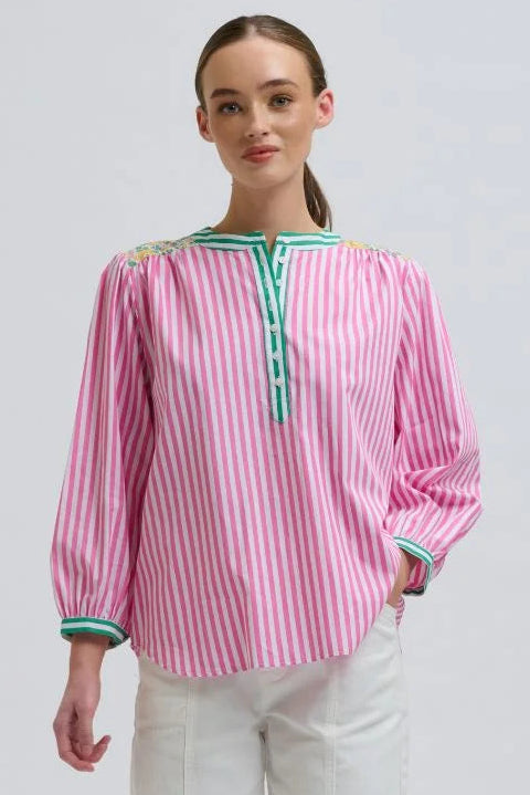 A woman modeling a pink and white vertically striped long-sleeve blouse with green trim at the collar and cuffs, paired with white wide-leg pants, against a light gray studio background.