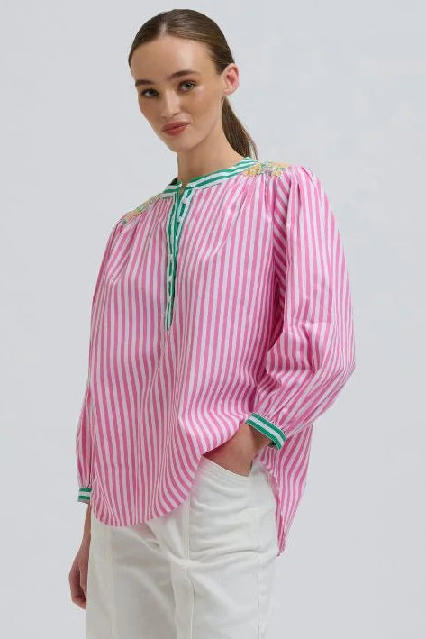 A woman modeling a pink and white vertically striped long-sleeve blouse with green trim at the collar and cuffs, paired with white wide-leg pants and beige woven sandals, against a light gray studio background.