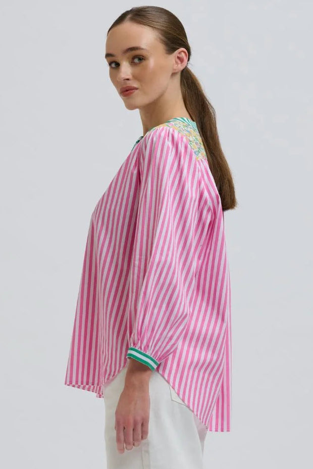 A woman modeling a pink and white vertically striped blouse with green and blue embroidered detailing on the shoulders and cuffs, paired with white trousers, against a light gray studio background.