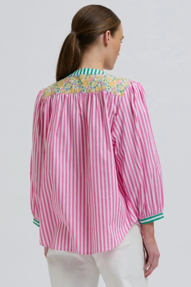 A woman shown from the back wearing a pink and white vertically striped blouse with a colorful floral embroidered yoke and white pants, against a light gray studio background.