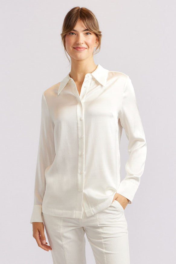 A woman standing against a white background, wearing a long-sleeved white silk shirt and matching pants, with dark shoes.